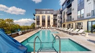 Located along Sibley Memorial Highway in Lilydale, Minnesota, this outdoor amenity space is designed to deliver a resort-style experience within a modern multi-story residential community. The centerpiece of the space is a generously sized swimming pool with crystal-clear blue water, accessible via stainless steel entry rails and complemented by a separate spa. The surrounding pool deck is finished in large-format concrete pavers, providing ample space for lounging and socializing.

Rows of cushioned chaise lounge chairs line the perimeter of the pool, offering residents abundant space for relaxation in the sun. Shaded cabana structures with striped cushions provide a comfortable retreat from direct sunlight. Additional seating areas are thoughtfully positioned throughout the deck, enhancing the social atmosphere of the space.

The building's exterior showcases a contemporary architectural design, combining light stone tile cladding, dark metal panel accents, and vertical board-and-batten siding. Private balconies extend from multiple floors of the building, many of which overlook the lush, wooded landscape beyond the property's perimeter railing. The natural treeline and open sky views lend the space a serene, nature-connected ambiance, making this outdoor amenity deck a standout feature of the Lilydale community.