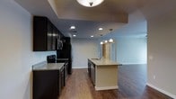 This single-floor residence offers a thoughtfully designed open-concept layout that seamlessly connects the kitchen and living areas, creating a spacious and inviting atmosphere. The kitchen is a standout feature, showcasing rich dark cabinetry that extends to the ceiling, providing ample storage while adding a bold, sophisticated aesthetic. Granite countertops with warm tones complement the cabinetry and extend across a central island, which is fitted with a sink and illuminated by elegant pendant lighting — making it ideal for both meal preparation and casual dining. Stainless steel appliances, including a built-in microwave, stove, and refrigerator, round out the fully equipped kitchen. Recessed lighting throughout ensures the space is well-lit, while the warm, dark hardwood flooring ties the entire open area together cohesively. The living area flows naturally from the kitchen, offering generous space with neutral light-toned walls that lend a clean, airy feel. A tray ceiling detail above the kitchen zone adds architectural dimension and visual interest. The overall ambiance of the space is modern and polished, balancing dark contrasting design elements with open, light-filled areas to create a comfortable and functional living environment.