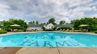 Located at 7200 Gorden Farms Parkway in Dublin, Ohio, this outdoor pool area serves as a central community amenity for the Gorden Farms neighborhood. The centerpiece is a generously sized swimming pool with a distinctive freeform curved design on one end, complemented by a classic rectangular section — offering a unique and visually appealing shape clearly branded with the Gorden Farms name on the pool floor. The pool is surrounded by an expansive concrete sun deck that provides ample space for relaxation and recreation. Scattered throughout the deck are lounge chairs and umbrella-shaded tables, offering residents a variety of options for sunbathing or socializing. Neatly trimmed hedges and mature trees frame the perimeter of the space, creating a sense of privacy and a lush, green backdrop. A white-sided clubhouse or pool house structure anchors one side of the area, featuring a charming pitched roofline and providing practical facilities for pool guests. The overall atmosphere is clean, well-maintained, and inviting, making it an ideal outdoor retreat during warmer months. The combination of thoughtful landscaping, generous deck space, and a well-appointed pool makes this a standout community feature in the Dublin, Ohio area.