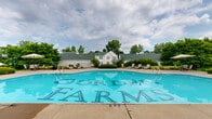 Located at 7200 Gorden Farms Parkway in Dublin, Ohio, this outdoor pool area serves as a central community amenity for the Gorden Farms neighborhood. The centerpiece is a generously sized swimming pool with a distinctive freeform curved design on one end, complemented by a classic rectangular section — offering a unique and visually appealing shape clearly branded with the Gorden Farms name on the pool floor. The pool is surrounded by an expansive concrete sun deck that provides ample space for relaxation and recreation. Scattered throughout the deck are lounge chairs and umbrella-shaded tables, offering residents a variety of options for sunbathing or socializing. Neatly trimmed hedges and mature trees frame the perimeter of the space, creating a sense of privacy and a lush, green backdrop. A white-sided clubhouse or pool house structure anchors one side of the area, featuring a charming pitched roofline and providing practical facilities for pool guests. The overall atmosphere is clean, well-maintained, and inviting, making it an ideal outdoor retreat during warmer months. The combination of thoughtful landscaping, generous deck space, and a well-appointed pool makes this a standout community feature in the Dublin, Ohio area.