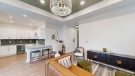 Located at 74 Callaway Dr NW in Calabash, North Carolina, this thoughtfully designed single-floor amenity space offers a compelling blend of modern aesthetics and functional versatility. The open lounge area serves as the heart of the space, anchored by a striking tray ceiling finished in deep olive green and adorned with an elegant beaded chandelier. Warm honey-toned hardwood flooring runs throughout, complementing the soft, neutral wall tones and curated furnishings that include a rich leather sofa, accent chairs, and tasteful artwork. Natural light filters in through multiple windows dressed with linen curtains, creating a welcoming and relaxed atmosphere. A dining nook near the entrance provides additional seating beneath the same distinctive tray ceiling detail. The kitchen is a standout feature, boasting crisp white shaker cabinetry, a deep green ceramic tile backsplash, light quartz countertops, a peninsula with bar seating, a built-in wine cooler, and matte black hardware throughout. Recessed lighting enhances every area of the kitchen. The dedicated exercise room offers a quiet, functional setting with recessed lighting, a ceiling fan, carpeted flooring, and natural light, equipped with free weights and a workout bench for fitness convenience.