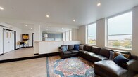This thoughtfully designed single-floor space offers a seamless open-concept layout that connects the living and kitchen areas, creating a fluid and functional environment. The living area is anchored by large, floor-to-ceiling windows that flood the space with natural light and frame sweeping elevated views of the surrounding cityscape and greenery beyond. Light hardwood flooring runs throughout, complementing the clean, neutral color palette of the walls and ceilings fitted with recessed lighting.

The kitchen is a standout feature, showcasing flat-front gray cabinetry stacked to near ceiling height for abundant storage, paired with crisp white subway tile backsplash and light stone countertops. A large central island with a butcher block top and white cabinetry provides additional prep space and casual seating, making it ideal for gathering. Stainless steel appliances, including a refrigerator, oven, and dishwasher, complete the well-appointed kitchen.

The living area offers a welcoming atmosphere, with a generously sized leather sectional sofa set over a richly patterned area rug. Multiple windows along the exterior wall ensure the space remains bright and connected to its urban surroundings. The overall ambiance is modern and inviting, combining clean architectural lines with warm, functional design elements throughout.
