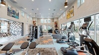 Located at 1230 Vintage Ln in Morgan Hill, California, this single-floor fitness center is a well-appointed community gym designed to support a comprehensive workout experience. The space is defined by its soaring double-height ceilings and whitewashed brick walls, which lend an open, airy feel throughout. Abundant natural light pours in through large floor-to-ceiling sliding glass doors and clerestory windows positioned along the upper walls, keeping the interior bright and energizing at all hours. Large-format sliding doors open directly to an outdoor terrace and pool area, offering refreshing views of the community amenity spaces during workouts. The cardio zone is thoroughly equipped with multiple treadmills, elliptical trainers, and stationary bikes, each featuring individual digital display screens. The strength training area includes a multi-station cable machine, a dedicated free weight rack, and a variety of resistance and functional fitness machines. Patterned carpet tiles in warm earth tones with orange accents define workout zones across the light-colored flooring, adding visual structure to the space. Pendant lighting and recessed fixtures provide even illumination throughout. Vibrant fitness-themed artwork adorns the walls, contributing to a motivating atmosphere. A water station is conveniently accessible near the exit. The overall design balances functionality with a modern, welcoming aesthetic.