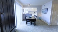This single-floor residence offers a cohesive, open layout that connects the dining area and kitchen in a practical and visually unified arrangement. The dining space is carpeted in a light, plush material that adds warmth underfoot and contributes to the overall comfortable atmosphere of the room. A dark-stained rectangular dining table with matching chairs serves as the central focal point, complemented by soft blue-gray walls that lend a calm, neutral tone throughout.

A pass-through opening connects the dining area directly to the kitchen, which features white cabinetry, white appliances, and a clean, functional design. This architectural detail enhances the sense of openness while maintaining a defined separation between spaces. The kitchen benefits from under-cabinet lighting that adds a subtle brightness to the work area.

Large vertical blinds frame a sliding glass door along one wall of the dining room, allowing for natural light and potential outdoor access. Flush-mounted ceiling lighting provides even illumination throughout the space. Decorative touches, including wall art and a vase with branches, add character without overwhelming the straightforward, well-organized layout of this single-story home.