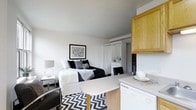 This well-appointed studio presents a thoughtfully designed single-level layout that maximizes every inch of available space. The open-plan interior combines the sleeping, living, and kitchen areas into one cohesive environment, creating a functional and inviting atmosphere.

The sleeping area anchors the main living space, featuring a full-sized bed dressed in crisp black-and-white bedding, complemented by a sleek bedside table with a modern lamp. A bold black-and-white chevron area rug defines the living zone and adds a striking graphic element to the room. Abstract wall art in a black frame contributes to the contemporary aesthetic.

A white wardrobe with mirrored doors provides generous storage and reflects light throughout the space, contributing to an airy, open feel. The white walls and smooth ceilings maintain a clean, minimalist backdrop throughout.

The kitchen area is efficiently tucked to one side, featuring warm honey-toned wood cabinetry overhead, a white countertop, and a dishwasher below. A compact dining table with a light wood base and white top seats two, serving as both a dining and workspace. The overall color palette of white, black, and natural wood tones creates a cohesive and modern living environment.