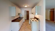 This single-floor unit presents a practical and well-organized layout across several distinct spaces. The galley-style kitchen is efficiently arranged, featuring white cabinetry with gold hardware, dark laminate countertops, a double-basin sink, and white appliances including a coil-top range and refrigerator. Tile flooring and a flush-mount ceiling light complete the kitchen, which connects to the rest of the unit through a wood-trimmed doorway.

The living area is generously proportioned, with neutral carpet, textured ceilings, and multiple windows that invite natural light and views of surrounding greenery. A wall-mounted air conditioning unit and baseboard heating provide year-round climate control. The open feel of the room offers versatile furniture arrangement possibilities.

The bedroom is a comfortable, quiet space finished with the same neutral carpet and textured ceiling. A window frames an outdoor tree view, while baseboard heating runs along the perimeter wall.

The bathroom is cleanly appointed with white tile flooring, a full tiled tub-shower surround, a toilet, and a vanity with a light countertop and white cabinetry. A wood-panel door and small window round out the space. Overall, the unit maintains a clean, understated aesthetic throughout.