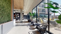 This contemporary amenity lounge is designed with an urban-industrial aesthetic, combining polished concrete floors, exposed ductwork, and warm wood ceiling accents to create a dynamic, loft-inspired atmosphere. The spacious open-plan layout is anchored by a full-size billiard table set atop a graphic area rug, serving as the social centerpiece of the room. Dark slate-blue accent walls are adorned with curated artwork and bold typographic murals, adding a creative energy throughout the space. A sleek built-in counter with light wood cabinetry, a white quartz surface, and a beverage refrigerator provides a convenient refreshment station, complemented by a geometric-patterned tile backsplash and a wall-mounted television. The lounge features multiple seating vignettes, including leather lounge chairs, bar-height counter seating, and casual dining tables, accommodating a range of social and work needs. Floor-to-ceiling black-framed glass walls and sliding doors flood the interior with natural light and offer direct access to a sunny outdoor terrace and resort-style pool deck lined with palm trees. A reception-style nook with a long desk, warm pendant lighting, and a wood-paneled ceiling provides a more refined gathering point. The seamless connection between interior and exterior living makes this lounge an inviting, versatile hub for community life.