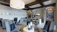 This single-floor space showcases a sophisticated blend of rustic and modern design. Exposed dark wood ceiling beams contrast beautifully against crisp white walls and light hardwood-style flooring, creating a warm yet refined ambiance throughout. A striking stone-clad archway serves as a dramatic focal point, framing a well-appointed bar area complete with pendant lighting, bar seating, and a wall-mounted display screen—ideal for social gatherings. The lounge area features plush grey upholstered seating, including accent chairs and sofas, arranged around a cylindrical white pedestal table adorned with decorative candles. Framed artwork punctuates the white-paneled walls, adding curated detail to the space. Toward the back, high-top tables and bar stools provide additional seating in a more casual setting. A large drum pendant light anchors the main lounge area, contributing to the layered lighting design. The overall atmosphere balances comfort and elegance, making the space well-suited for relaxed socializing or more structured gatherings. The thoughtful combination of textures—stone, wood, fabric, and metal—gives the interior a polished, boutique-style character.