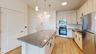 Located at 320 Fisher St in Charlottesville, VA, this single-floor unit offers a well-designed, contemporary interior with a clean, neutral color palette throughout. The kitchen serves as a central highlight, featuring light wood cabinetry with matte black hardware, granite countertops, a subway tile backsplash, and a full suite of stainless steel appliances including a gas range, built-in microwave, dishwasher, and refrigerator. A large kitchen island with a matching granite surface and pendant lighting overhead provides additional workspace and a natural gathering point that opens toward the living and dining areas.

The living area is finished with light wood-tone flooring, offering a bright and airy atmosphere, and includes direct access to an outdoor balcony through a glass door framed by large windows that bring in ample natural light and greenery views. A separate dining space adjacent to the kitchen adds functional versatility.

The bedroom is carpeted for comfort, equipped with a ceiling fan, and benefits from natural light through a well-positioned window. It connects directly to a spacious walk-in closet with wire shelving along multiple walls. The en-suite bathroom is generously proportioned, featuring a dual-sink vanity with granite countertops, a glass-enclosed shower, and hardwood-style flooring, contributing to an overall polished and functional living environment.
