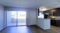 This single-floor residence offers a clean, functional layout with a welcoming open-plan design. The living area is finished with rich, dark hardwood-style flooring that runs throughout, lending warmth and continuity to the space. A large sliding glass door fitted with vertical blinds fills the room with natural light and provides direct access to an outdoor area, creating a bright and airy ambiance.

Adjacent to the living area is a compact kitchen, thoughtfully positioned to maintain an open connection to the main living space. The kitchen features dark espresso-toned cabinetry that contrasts with a light-colored countertop and a tiled backsplash, lending a modern, two-toned aesthetic. A black refrigerator anchors the space, and flush-mount ceiling lighting ensures the kitchen remains well-lit.

A white front entry door with brushed hardware serves as the focal point of the main wall, establishing a clear sense of arrival and separation between the living area and the entry point. The neutral wall tones throughout complement the darker flooring and cabinetry, creating a balanced and cohesive interior palette. Overall, the space is designed for practical, everyday living with a modern residential character.
