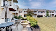 This residential community offers a welcoming and well-appointed living environment set amidst lush, manicured grounds. The multi-story buildings feature a classic architectural style with neutral siding, burgundy accent shutters, and pitched rooflines that lend a warm, traditional character to the property. Each unit is equipped with private balconies or ground-level patios, providing residents with personal outdoor spaces to enjoy the surrounding scenery.

The landscaping is a standout feature of the community, with neatly trimmed hedgerows, mature trees, vibrant flowering planters, and expansive green lawns that create a serene, park-like atmosphere throughout the grounds. Decorative stone urns with seasonal blooms serve as elegant focal points along the walking paths.

Outdoor communal areas include a furnished patio with table and chair seating, ideal for relaxing or socializing in a peaceful setting. The well-maintained pathways connect the buildings and green spaces, encouraging an active and connected community lifestyle. Ground-floor units feature sliding glass doors that open directly to the patio areas, seamlessly blending indoor and outdoor living. Overall, the property presents a tranquil and thoughtfully designed environment with strong curb appeal and inviting common spaces.