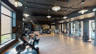 This stylish fitness center combines industrial design with a high-end gym aesthetic across a single, open floor plan. The space features a dark, dramatic ceiling with exposed ductwork and black structural beams, contrasted by warm pendant lighting and track lighting throughout. Light hardwood flooring covers the main workout area, transitioning to dark rubber flooring in the strength and weight training zones.

The cardio area is well-appointed with Peloton spin bikes, treadmills, elliptical machines, stair climbers, and rowing machines. Treadmills are positioned near large floor-to-ceiling windows that flood the space with natural light and offer views of the surrounding greenery. A dedicated strength training zone features a Smith machine, a functional training rig with TRX straps and medicine balls, and a full rack of dumbbells. Vertical LED light strips and full-length mirrors in the strength area add to the sleek, modern atmosphere.

A textured stone accent wall anchors the free weights section, providing visual warmth against the matte dark walls. The open layout ensures ample space between equipment, allowing for comfortable movement throughout. The combination of natural light, thoughtful lighting design, and premium equipment makes this a comprehensive and inviting fitness facility.