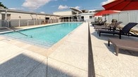 This outdoor amenity space centers around a generously sized swimming pool with crystal-clear blue water, set within a bright, open-air environment under expansive skies. The pool area is fully enclosed by a white aluminum security fence, providing both safety and a clean, modern aesthetic. A wide, light-colored aggregate concrete deck surrounds the pool on multiple sides, offering ample space for movement and relaxation. Linear drainage channels are integrated along the deck's edges, reflecting thoughtful infrastructure design.

Adjacent to the pool, a dedicated sunbathing area features wicker-style chaise lounge chairs accompanied by large red shade umbrellas, providing comfortable options for lounging in the sun or shade. The surrounding landscape incorporates decorative river rock ground cover along the fence line, adding a low-maintenance yet polished finishing touch alongside patches of green lawn visible beyond the perimeter.

Covered pergola-style walkways with arched canopies line portions of the enclosure, providing shaded access routes around the facility. The broader building complex, visible in the background, features a flat-roofed, single-story design with large windows and additional covered overhangs. The overall atmosphere is clean, functional, and resort-like, designed for comfortable outdoor recreation and leisure.
