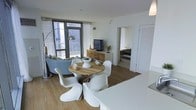 This single-floor space showcases a thoughtfully designed open-plan layout that seamlessly connects the kitchen, dining, and living areas. The kitchen features sleek white countertops that flow naturally into the dining space, where a rustic wooden round table is paired with iconic white Panton-style chairs, creating a striking contrast of materials. A cozy seating area with a curved sofa and an arc floor lamp adds a relaxed ambiance to the living zone. Floor-to-ceiling windows with sheer curtains flood the interior with abundant natural light while framing impressive views of the surrounding urban skyline. The flooring throughout is warm wood-tone laminate, adding a grounding element to the predominantly white palette. A wooden media console houses a flat-screen television, providing functional storage while complementing the natural tones of the dining table. A doorway visible from the main living area leads to a bedroom, offering a sense of privacy while maintaining the open feel of the overall layout. The combination of white walls, modern furnishings, and natural light creates a clean, airy, and contemporary atmosphere throughout the space.
