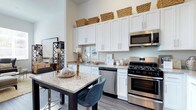 The S3 studio is a thoughtfully designed single-floor residence that makes a strong first impression with its open, airy layout and modern finishes. The kitchen serves as a central highlight of the space, showcasing floor-to-ceiling white shaker cabinets that maximize storage capacity and draw the eye upward, emphasizing the notably tall ceiling height. Stainless steel appliances — including a gas range, built-in microwave, and dishwasher — provide a sleek, contemporary aesthetic alongside functionality. The granite countertops feature a light, speckled finish that complements the clean white cabinetry, while a kitchen island with bar seating offers an ideal spot for casual dining or entertaining. Light wood-style plank flooring runs throughout, adding warmth and continuity to the space. The overall ambiance is bright and modern, with a neutral color palette that creates a versatile canvas. Every detail, from the under-cabinet hardware to the open sightlines of the layout, reflects a commitment to both style and practical everyday living in this well-crafted studio.