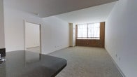 This single-floor space presents an open, functional layout with a seamless flow between its main living areas. The living room is finished with neutral-toned carpeting that adds warmth underfoot, while crisp white walls create a bright and airy atmosphere throughout. A standout architectural feature is the exposed brick wall accent surrounding a large window, which allows natural light to pour into the space and adds a touch of industrial character to the otherwise clean, modern interior. The window itself offers a view outward, enhancing the sense of openness. A dark granite countertop is visible in the foreground, suggesting a kitchen or bar area that transitions naturally into the main living space. Additional rooms are accessible through open doorways, hinting at a well-connected floor plan. The smooth white ceilings and neutral color palette throughout provide a versatile backdrop suitable for a variety of uses and interior styles. Overall, the space combines classic and contemporary elements — exposed brick, granite surfaces, and soft carpeting — to create a balanced, inviting environment.