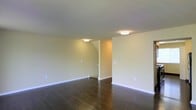 This space presents a straightforward, functional layout spread across its levels, connected by a carpeted staircase with white-painted risers and a dark wood handrail running along one side. The stairs are finished with a dark brown, textured carpet that continues throughout the interior, lending a consistent and cohesive feel underfoot.

The main living area is open and generously sized, with smooth, light-colored walls that create a clean, neutral backdrop. Two open doorways lead to adjoining rooms, both of which benefit from natural light provided by windows that frame views of greenery outside. One of the connecting rooms features a wooden dresser and continues the warm carpeted flooring, suggesting use as a bedroom or personal space. The other adjoining room is fitted with a ceiling-mounted light fixture and also receives natural light, indicating it serves as an additional functional room.

Throughout the space, the design is minimal and practical, with white painted walls, standard ceiling heights, and carpeted floors providing a quiet, comfortable atmosphere. The open sightlines between rooms enhance the sense of connectivity and flow within the home.