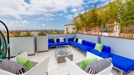This rooftop terrace offers a well-appointed outdoor retreat designed for both relaxation and social gatherings. The space is finished with light marble-style tile flooring that lends a clean, sophisticated aesthetic throughout. Large rectangular planters filled with bamboo, flowering shrubs, and ornamental grasses are thoughtfully positioned to provide natural privacy screening while adding vibrant greenery to the environment.

The terrace is divided into distinct functional zones. A lounge area features a generous sectional sofa with bold blue cushions and accent pillows in green and black-and-white patterns, arranged around a central fire pit table with a glass surround and river rock fill — an ideal setting for evening gatherings. A separate dining zone includes black tables paired with bright green modern chairs, offering a lively contrast to the neutral palette of the surrounding architecture.

Additional seating options, including hanging egg-style chairs, provide casual nooks for relaxation. The open layout allows for unobstructed panoramic views of the surrounding cityscape and sky, making the most of its elevated position. The combination of curated furnishings, layered greenery, and city views creates an inviting and functional outdoor amenity space suited for a wide range of uses.