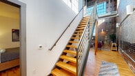 Located at 144 4th St in Burlington, NC, this two-story, 3-bedroom, 2-bathroom space showcases a striking industrial character defined by soaring exposed brick walls, large factory-style windows, and warm hardwood flooring throughout. The main floor hosts the expansive living area, where dramatic double-height brick walls serve as a bold architectural backdrop, flooding the space with natural light through oversized windows. The kitchen features rich dark wood cabinetry, granite countertops, stainless steel appliances including a full refrigerator, range, microwave, and dishwasher, complemented by exposed ductwork overhead that reinforces the industrial aesthetic. A sleek open-tread staircase with steel railings and warm wood steps connects the main floor to the upper level, where additional bedrooms are located. The spacious primary bedroom offers hardwood flooring and a generous closet with built-in shelving and hanging rods. The bathrooms are finished with granite-topped dark wood vanities, large mirrors, and clean white fixtures. Throughout the space, the interplay of raw brick, white walls, natural wood, and steel creates a cohesive urban-industrial ambiance that is both functional and visually compelling.
