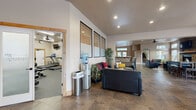 This two-floor community clubhouse offers a well-rounded selection of amenities designed for comfort, recreation, and productivity. The entrance foyer makes a strong first impression with striking stacked-stone accent walls, decorative mountain wall art, and a welcoming seating nook with leather chairs and a round coffee table set on a circular area rug.

The main lounge area features plush black leather sofas accented with bold red and yellow throw pillows, arranged around a rustic wood coffee table on a textured area rug. Large windows with wood-trimmed blinds bring in ample natural light throughout.

Adjacent to the lounge is a fully equipped kitchen with light maple cabinetry, dark granite countertops, stainless steel appliances, and a standout stone-clad breakfast bar with seating — ideal for gatherings and events.

The spacious game room is bathed in natural light from a sweeping bank of large windows and features a full-size pool table, a stone fireplace, and a long bar-height counter with seating along one wall.

A dedicated business center includes desktop computers at a communal high-top table and opens to the outdoors via French doors. The fitness center is well-appointed with cardio machines, cable weight equipment, dumbbells, medicine balls, rubber flooring, wall mirrors, and direct outdoor access.