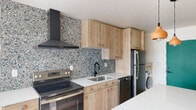 This thoughtfully designed single-floor space showcases a cohesive modern aesthetic throughout. The kitchen is a standout feature, boasting light wood cabinetry with distinctive rounded hardware, crisp white quartz countertops, and a striking terrazzo-style backsplash that extends dramatically up the wall. A matte black range hood anchors the cooking zone above a stainless steel electric range, complemented by a black undermount sink with a matching matte black faucet. A central kitchen island with pendant lighting overhead provides additional prep and seating space, while stainless steel appliances, including a refrigerator and dishwasher, complete the culinary setup. In-unit laundry is neatly tucked beneath cabinetry with a dedicated counter surface, offering everyday convenience. The living area benefits from natural light streaming through large windows, with light hardwood-style flooring flowing throughout and contributing to the home's bright, airy ambiance. The bathroom is elegantly finished with warm terracotta-toned wall tiles, a corner glass-enclosed shower with matte black fixtures, a wood-vanity with a white basin, and an arched mirror. A teal entry door adds a bold pop of color, lending a distinctive and welcoming character to the space.