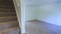 Located at 1844 Forest Village Lane in Columbus, Ohio, this two-story home presents a straightforward and functional layout designed for comfortable everyday living. The interior features neutral white walls throughout, creating a bright and clean canvas that can easily be personalized to suit any style. Plush wall-to-wall carpeting runs through the main living areas, adding warmth and comfort underfoot. A carpeted staircase with a classic handrail connects the two floors, positioned conveniently to allow for an open and accessible flow between levels. The main floor living area is generously proportioned, offering ample space for a variety of furniture arrangements and uses. The open wall surfaces and simple architectural lines give the home a clean, uncluttered feel. Floor vents are integrated into the flooring, indicating a central forced-air system for year-round climate control. The overall ambiance of the home is calm and move-in ready, with its neutral palette and well-maintained finishes making it a versatile space suitable for a range of lifestyles.