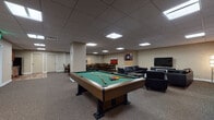 Located at 1717 E Ninth St in Cleveland, Ohio, this single-floor amenity space is designed to serve as a well-rounded gathering and recreation hub. The layout seamlessly integrates multiple functional zones within an open, cohesive environment finished with neutral-toned carpet and a suspended tile ceiling fitted with recessed panel lighting throughout.

At the heart of the space sits a full-size billiards table, serving as the centerpiece of the game area. Nearby, a dartboard mounted on the wall adds an additional recreational element. Adjacent to the game area is a comfortable lounge zone furnished with sleek black leather sofas and armchairs arranged around glass coffee tables, with a large flat-screen television and contemporary artwork adorning the walls.

The dining and gathering areas feature multiple solid wood dining tables with upholstered ladder-back chairs, capable of accommodating a sizeable group. Bold, colorful canvas artwork punctuates the neutral walls, adding visual interest and energy to the space. A kitchenette area with cabinetry and appliances is also present, supporting catering and refreshment needs.

The overall ambiance is modern and functional, balancing recreational entertainment with comfortable social seating, making it well-suited for community events, casual gatherings, and leisure activities.