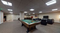 Located at 1717 E Ninth St in Cleveland, Ohio, this single-floor amenity space is designed to serve as a well-rounded gathering and recreation hub. The layout seamlessly integrates multiple functional zones within an open, cohesive environment finished with neutral-toned carpet and a suspended tile ceiling fitted with recessed panel lighting throughout.

At the heart of the space sits a full-size billiards table, serving as the centerpiece of the game area. Nearby, a dartboard mounted on the wall adds an additional recreational element. Adjacent to the game area is a comfortable lounge zone furnished with sleek black leather sofas and armchairs arranged around glass coffee tables, with a large flat-screen television and contemporary artwork adorning the walls.

The dining and gathering areas feature multiple solid wood dining tables with upholstered ladder-back chairs, capable of accommodating a sizeable group. Bold, colorful canvas artwork punctuates the neutral walls, adding visual interest and energy to the space. A kitchenette area with cabinetry and appliances is also present, supporting catering and refreshment needs.

The overall ambiance is modern and functional, balancing recreational entertainment with comfortable social seating, making it well-suited for community events, casual gatherings, and leisure activities.
