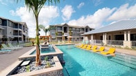 The outdoor amenity area of this residential community centers around a stunning resort-style swimming pool, designed to evoke a vacation atmosphere. The pool features crystal-clear turquoise water, complemented by a raised spa with elegant water features that cascade into the main pool below. The surrounding deck is generously sized, offering ample space for relaxation, and is furnished with bright yellow chaise lounges that add a vibrant, cheerful contrast to the blue water.

Tropical landscaping enhances the ambiance, with tall palm trees and decorative rock gardens framing the pool area and lending a lush, resort-like feel. A covered cabana structure sits adjacent to the pool, providing shaded seating for those seeking respite from the sun. A pergola can also be seen in the background, adding architectural interest to the outdoor space.

The multi-story residential building looms as a backdrop, featuring craftsman-inspired architectural details including dormer accents and covered balconies. The entire pool area is enclosed by a wrought-iron style fence, ensuring both safety and security. Open skies and natural sunlight flood the space, creating an inviting, vibrant outdoor retreat.