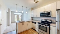 Located at 1399 Walton Way in Augusta, Georgia, this single-floor unit offers a clean, modern aesthetic throughout its well-appointed layout. The kitchen serves as a functional centerpiece, featuring stainless steel appliances including a refrigerator, electric range, built-in microwave, and dishwasher. White cabinetry with sleek hardware provides ample storage, while neutral countertops and warm wood-look flooring create a cohesive, contemporary feel. Pendant lighting hangs above the peninsula, which includes a built-in sink and additional cabinet storage below, offering both workspace and a natural division between the kitchen and the adjoining living area. The living space benefits from natural light streaming through glass-panel doors that open to a private balcony. From the balcony, views extend over a landscaped courtyard with seating areas, a pool, and the surrounding neighborhood. The full bathroom is finished with the same wood-look flooring, a white vanity with a vessel sink and dark countertop, a large mirror, and a tiled shower-tub combination. The overall design of the unit is consistent and thoughtfully executed, with a neutral color palette, clean lines, and modern finishes that create a bright, functional, and inviting living environment.