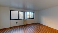 This single-floor residential unit offers a clean, straightforward layout designed for comfortable everyday living. The living area is a standout feature, characterized by warm hardwood-style flooring that extends throughout the space, crisp white walls, and a generous bank of large windows that invite abundant natural light and provide views of the surrounding neighborhood. A wall-mounted heating unit is positioned beneath the windows, ensuring year-round comfort. The open layout of the living area offers considerable flexibility for furniture arrangement and personal customization. Adjacent to the living space, the kitchen is well-appointed with warm maple-toned cabinetry — both upper and lower — providing ample storage. The countertops feature a granite-patterned surface complemented by a stainless steel sink, while a white gas range completes the functional cooking setup. The bedroom continues the consistent hardwood-style flooring and white walls, creating a calm and neutral environment conducive to rest. The bathroom is neatly appointed with a wood-toned vanity, a mirrored medicine cabinet, classic white tile flooring, and a combined shower and bathtub. Overall, the unit presents a practical and cohesive living environment with consistent finishes throughout.