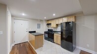 This single-floor unit offers a thoughtfully arranged layout that maximizes its compact footprint. The kitchen serves as the centerpiece, featuring light wood cabinetry with raised-panel doors, dark granite countertops, and a suite of black appliances including a gas range with oven, a full-size refrigerator, and a built-in sink. A peninsula with matching granite provides additional counter space and separates the kitchen from the adjacent living area. Recessed lighting illuminates both zones, and the transition between white ceramic tile in the kitchen and warm hardwood flooring in the living area clearly delineates each space. The living area is open and versatile, with neutral beige walls, recessed lighting, and a wall-mounted split-unit air conditioner for climate control. Small windows positioned near the ceiling allow natural light to filter in. A closet and additional storage space are accessible within the living area. The bathroom is clean and functional, finished with beige floor tile and white wall tile surrounding a full tub-shower combination. A toilet, vanity, and an over-toilet storage cabinet complete the space efficiently.