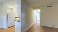 This single-floor space is designed with a straightforward, functional layout that emphasizes clean lines and a neutral aesthetic throughout. The interior features light-toned hardwood flooring that runs continuously from the kitchen area through the hallway and into the rooms beyond, creating a sense of cohesion and flow across the unit.

A partial wall with a dark countertop defines the kitchen area to one side, offering a semi-open concept that separates the cooking space without fully closing it off from the corridor. White cabinetry is visible, contributing to a bright and tidy kitchen environment.

The central hallway is wide and unobstructed, with smooth neutral-toned walls, white baseboards, and flush ceiling lighting that maintains a clean, understated look. A door at the far end opens into a room filled with natural light from a window, suggesting a bedroom or den that benefits from good daylighting. The overall ambiance of the space is calm and minimal, with a practical design suited to everyday living. The consistent use of white trim, neutral wall colors, and warm wood flooring ties the space together cohesively.