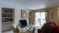 This single-floor residence offers a warm and inviting living space designed for comfortable everyday living. The living room serves as the heart of the home, anchored by a classic fireplace with a dark surround and white mantel that adds a timeless focal point to the room. A built-in shelving nook beside the fireplace provides practical display and storage space. The textured ceiling adds architectural character throughout, while neutral-toned walls create a bright and versatile backdrop. Natural light fills the room through a set of sliding glass doors dressed with flowing curtains, which open directly onto an outdoor patio area, offering a seamless transition between indoor and outdoor living. The patio is visible beyond the glass doors, featuring outdoor seating surrounded by greenery, suggesting a pleasant area for relaxation. The living area comfortably accommodates a seating arrangement centered around a glass-top coffee table, anchored by a patterned area rug that adds texture and warmth underfoot. Overall, the space presents a cozy, functional layout with classic design details that create an approachable and livable ambiance.