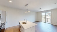 This single-floor residence showcases a bright, contemporary design with a thoughtful open-concept layout. The kitchen serves as the centerpiece, anchored by a generous white island with quartz countertops, an undermount sink, and a sleek linear pendant light overhead. White shaker-style cabinetry lines the walls alongside stainless steel appliances, including a refrigerator, range, and microwave. Light wood-style flooring flows seamlessly from the kitchen into the living area, which opens directly onto a private balcony through sliding glass doors, offering neighborhood views.

The bedrooms feature soft carpet flooring and large windows that bring in ample natural light, with views of the surrounding community. The neutral cream-toned walls throughout the unit create a clean, versatile backdrop. The bathroom is well-appointed with a wide quartz vanity, an undermount sink with chrome fixtures, a large mirror, and a glass-enclosed shower, all finished with light wood-style flooring consistent with the unit's modern aesthetic. Overall, the space maintains a cohesive, minimalist palette with quality finishes throughout.