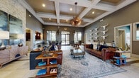 This single-story community clubhouse blends Western-inspired aesthetics with contemporary sophistication throughout its thoughtfully designed spaces. The grand entrance greets visitors with soaring ceilings, large-format tile flooring accented by wood-inlay detailing, and a stately solid wood door with decorative glass panels set against an exposed stone surround. Just beyond, a spacious leasing office features a warm wood reception desk, pendant lighting with Edison-style bulbs, a stacked stone accent column, and a patterned accent wall that adds visual depth. A dedicated consultation seating area with high-top tables and modern chairs completes the workspace. The expansive lounge is the heart of the clubhouse, showcasing dramatic coffered ceilings with wood-paneled insets and a striking sputnik chandelier. Rich leather Chesterfield sofas are arranged atop a layered area rug, while a gallery of sculptural white longhorn skulls commands an entire accent wall, reinforcing the space's bold Western character. Large floor-to-ceiling windows and glass doors flood the interior with natural light and provide direct views of and access to the outdoor pool area. A dining alcove near the pool exit features round tables, upholstered chairs, and tiered pendant fixtures, creating a refined yet relaxed setting for gathering and socializing.