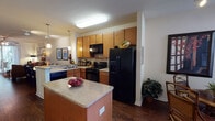 This single-story residence at 8450 Gate Pkwy W in Jacksonville, Florida offers a well-designed, open layout that connects the main living areas seamlessly. The kitchen is a functional centerpiece, featuring warm honey-toned wood cabinetry, granite-style countertops, black appliances including a side-by-side refrigerator, electric range, and dishwasher, all complemented by a central island that provides additional prep space and casual dining potential. Pendant lighting above the island adds a modern touch, while the rich hardwood-style flooring flows throughout the main living areas. Adjacent to the kitchen, a dedicated dining area accommodates a full dining set and benefits from natural light through large windows. The living room is generously proportioned, with sliding glass doors that allow ample natural light and access to an outdoor space. Ceiling fans are present in the bedrooms and living areas, enhancing comfort year-round. The bedrooms feature plush carpeting, multiple windows with blinds for privacy, and ample room for furnishings. The bathroom is well-appointed with warm wood vanity cabinetry, a wide countertop, a large mirror with overhead lighting, and a tub-shower combination with tile surround. Overall, the space presents a comfortable and cohesive interior with a warm, neutral palette throughout.