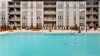 Located at 3389 Endeavor Ln in Vestavia Hills, Alabama, this outdoor amenity space centers around a generously sized resort-style swimming pool that serves as the focal point of a well-designed courtyard setting. The pool features clean, light-toned tile work and is surrounded by a broad, paved deck outfitted with rows of lounge chairs, offering ample space for relaxation and sunbathing.

The surrounding multi-story building presents a modern architectural design, characterized by a gray and neutral-toned exterior with horizontal siding, large windows, and private balconies that overlook the pool area. Tudor-inspired gabled rooflines add a distinctive architectural accent to the building's facade, blending contemporary and traditional design elements.

A covered gazebo with a brick base and pitched roof anchors one corner of the pool deck, providing shaded seating and a gathering space for residents. Additional seating areas are positioned around the perimeter, enhancing the social and recreational appeal of the outdoor space. Low ornamental plantings and landscaped beds line the edges of the courtyard, softening the hardscape and adding greenery to the environment.

The layout of the courtyard is thoughtfully designed to maximize open space while maintaining a cohesive aesthetic that complements the architecture of the surrounding building. The space presents a polished, community-oriented outdoor amenity with strong visual appeal.