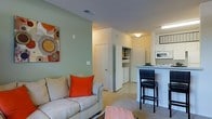 Located at 9525 US-78 Hwy in Ladson, South Carolina, this single-floor residence offers a well-designed open-concept layout that seamlessly connects the main living areas. The living room is a welcoming space, highlighted by a soft color palette with a sage green accent wall that adds depth and character. The open design flows naturally into the kitchen, which features a raised breakfast bar with seating — perfect for casual dining or entertaining. The kitchen is equipped with white appliances, including a built-in microwave and oven, set against clean white cabinetry and countertops that keep the space feeling bright and functional. A dedicated laundry area is conveniently tucked within the unit, providing added practicality. Storage is thoughtfully incorporated into the layout, with a built-in shelving nook offering organized space for everyday essentials. The flooring transitions between carpet in the living areas and hard flooring near the kitchen and utility zones, defining each space while maintaining an open, cohesive feel. Recessed and overhead lighting throughout ensures the interior remains well-lit at all times. The overall ambiance is clean, practical, and inviting, making this a comfortable and efficiently organized living space in the Ladson community.