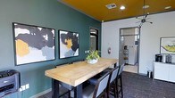 This single-floor space makes a strong visual impression through its carefully curated interior design. The dining or meeting area serves as a focal point, anchoring the room with a substantial natural wood communal table surrounded by sleek, dark-framed chairs upholstered in cool gray. The combination of materials — raw wood against industrial metal — strikes a balance between warmth and contemporary edge.

The color scheme is notably bold: deep teal walls contrast dramatically with a rich mustard-yellow ceiling, creating an energetic yet cohesive atmosphere. Recessed lighting in the ceiling provides focused illumination, complementing the overall design. A set of three framed abstract artworks adorns the teal accent wall, featuring blacks, whites, and golden yellows that echo the room's palette and reinforce its modern aesthetic.

A small potted plant centered on the table adds a natural, welcoming touch. Beyond the dining area, a hallway leads to additional spaces, hinting at further utility. A shelving unit and additional furnishings are visible through the corridor, suggesting the space extends into functional support areas. Overall, the design feels intentional, modern, and well-suited for communal use.