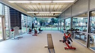 This fitness center offers a comprehensive workout environment thoughtfully organized into distinct training zones. The facility is divided into two primary areas: a bright cardio and cycling zone, and a dedicated strength training area, each designed to support a full range of exercise activities.

The cardio zone features an array of spinning bikes, rowing machines, elliptical trainers, and treadmills arranged along expansive floor-to-ceiling glass walls. These large-panel windows and sliding glass doors flood the space with natural light and provide open, unobstructed views of adjacent green courts and mature trees, creating an energizing atmosphere during workouts.

The strength training area is outfitted with a comprehensive selection of cable and selectorized machines, free weights, a full dumbbell rack, and flat benches. Large mirrors line the walls, enhancing the sense of space and allowing users to monitor their form. The flooring transitions between speckled rubber and a distinctive blue rubberized surface, clearly delineating the training zones.

Overhead, an exposed structural ceiling with long-span metal decking houses ceiling fans and linear fluorescent lighting, contributing to the industrial-modern aesthetic of the facility. The space also features decorative artwork near the entrance, adding a welcoming touch. The overall ambiance is bright, open, and functional, making it a well-rounded fitness destination.