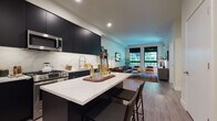 This single-floor residence showcases a thoughtfully designed open-concept layout that seamlessly connects the kitchen and living areas. The kitchen is outfitted with floor-to-ceiling matte dark cabinetry, a marble-style backsplash, stainless steel appliances including a French-door refrigerator, gas range, built-in microwave, and dishwasher. A central island with a white quartz countertop provides additional prep space and casual seating with bar-height chairs. Light wood-tone flooring runs throughout, creating a warm contrast against the dark cabinetry.

The living area extends toward a bank of large, grid-paned windows that flood the space with natural light and offer views of lush greenery outdoors. Recessed lighting and clean white walls complement the open, airy ambiance. The bedroom continues the same window style, enhancing the sense of light and connection to the outdoors.

The bathroom features a floating dark vanity with a white countertop, an integrated sink, and a large mirror, maintaining the consistent modern aesthetic found throughout the unit. Overall, the space balances contemporary design with warm, livable details.