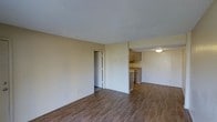 This single-floor space presents a straightforward, functional layout designed to maximize usability. The main living area is open and unobstructed, finished with warm wood-style laminate flooring that runs consistently throughout, adding a cohesive and clean aesthetic underfoot. The walls are painted in a neutral beige tone, contributing to a bright and understated atmosphere. The textured white ceiling adds a subtle decorative element while maintaining a modest overhead profile. A doorway off the main living area leads to an additional room, offering separation between living and private spaces. The kitchen is partially open to the living area, separated by a half-wall that creates a defined yet connected cooking space. It features light wood cabinetry providing ample storage, along with countertop workspace and standard appliances. A flush-mount ceiling light fixture illuminates the kitchen area. Overall, the space presents a practical and neutral interior that offers a functional foundation, with consistent flooring and a calm, muted color palette throughout.