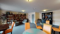 This single-floor community space serves as both a social gathering area and a library lounge, thoughtfully designed to encourage relaxation and interaction. The room is finished with soft blue carpeting throughout and features crown molding along the ceiling, lending a polished, comfortable character to the interior.

The library corner is well-appointed with tall wooden bookshelves filled with a wide selection of books, complemented by a pair of upholstered armchairs — ideal for quiet reading. Adjacent to this area stands a striking dark-painted display cabinet with lower storage drawers and open shelving above, showcasing decorative items including a globe, ship models, and nautical accents that add a classic, distinguished touch.

The dining and gathering area features multiple round glass-top tables surrounded by vibrant orange leather chairs, creating a bright and inviting setting for casual conversation or group activities. A framed mirror and wall sconces add decorative warmth to the walls in this section.

Large windows along one side of the room flood the space with natural light and offer views of the surrounding outdoor area, while a glass door provides convenient direct access to the exterior. The overall ambiance is warm and communal, balancing functionality with a homey, welcoming aesthetic suited for everyday use.