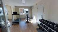 This single-floor fitness room offers a practical and well-organized space for exercise and wellness. The room features hardwood-style flooring throughout, lending a clean and polished look to the workout environment. Natural light streams in through a window that overlooks an outdoor area, creating a bright and inviting atmosphere. The space is equipped with a treadmill and an elliptical trainer, providing options for cardiovascular workouts, while a dedicated dumbbell rack stocked with a range of free weights caters to strength training needs. A wall-mounted unit provides climate control, ensuring comfort during workouts regardless of the season. Baseboard heating along the perimeter adds an additional layer of temperature regulation. The neutral, light-toned walls contribute to an open and airy feel, while the overall layout is efficiently arranged to maximize usable floor space. A secure entry door provides easy access. This fitness room is well-suited for residents or guests seeking a convenient, on-site exercise facility with essential equipment for a complete workout routine.