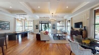 This elegantly designed single-floor community lounge or clubhouse space exudes a refined, upscale ambiance throughout. Rich, warm hardwood floors flow seamlessly across the entire space, complementing the crisp white coffered and tray ceilings adorned with detailed crown molding. Generous natural light pours in through large, black-framed floor-to-ceiling windows and glass doors, creating a bright and welcoming atmosphere with views of the surrounding exterior.

The main gathering area is anchored by a plush seating arrangement centered around a coffee table, beneath a stylish circular chandelier. Adjacent to this is a dedicated billiards area, offering a social and recreational focal point. A separate seating nook features a round glass-top table surrounded by upholstered chairs, ideal for intimate gatherings.

The space also includes a well-appointed wet bar with rich dark wood cabinetry, granite countertops, bar seating, a beverage refrigerator, and a wall-mounted TV framed by built-in shelving — perfect for entertaining. A secondary TV lounge area features a wall-mounted flat-screen above a decorative dark wood media console, flanked by large windows with drapery. Throughout, carefully chosen furnishings, artwork, and décor contribute to a polished, community-focused environment.