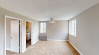 This single-floor unit offers a practical and comfortable layout designed to maximize livable space. The living area is finished with plush wall-to-wall carpeting and neutral gray walls, creating a warm and inviting atmosphere. A ceiling fan with a light fixture provides both air circulation and overhead lighting, while a classic cast-iron radiator ensures efficient heating. Multiple windows throughout the unit allow natural light to flow in, offering views of the surrounding neighborhood.

An additional carpeted room, also equipped with a radiator and a large window, serves well as a bedroom or flexible-use space. The compact kitchen is fitted with rich honey-toned wood cabinetry — both upper and lower — along with a stainless steel sink, a white electric range, and a full-size refrigerator. Dark hardwood-style flooring in the kitchen adds contrast to the lighter finishes elsewhere in the unit.

The bathroom features tile flooring in warm earth tones, a full-size soaking tub with an overhead shower, and clean white fixtures throughout. The overall layout, as seen from the dollhouse view, is efficiently organized, with rooms flowing naturally from one to the next, making this a functional and comfortable living space.