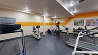 Located at 28500 Mission Blvd in Hayward, California, this fitness center offers a fully equipped workout space contained within a single room. The interior is defined by a bold design aesthetic, with striking orange and yellow horizontal stripe accents running along cream-colored walls, giving the space a vibrant, energetic atmosphere.

The room is outfitted with a variety of cardio machines, including multiple treadmills, an elliptical trainer, a stationary bike, and a stair climber, providing a well-rounded selection of aerobic exercise options. A dedicated free weight area features a multi-tier dumbbell rack, catering to strength training needs. A stability ball and additional accessories round out the available equipment.

A large wall-mounted mirror spans one side of the room, visually expanding the space and allowing users to monitor their form during workouts. The dark rubber flooring provides durability and traction underfoot. Recessed and flush-mounted ceiling lights ensure consistent, even illumination throughout. Ventilation is supported by ceiling-mounted air vents, maintaining a comfortable environment during exercise. A single entry door with a window provides access, and a fire extinguisher is mounted on the wall for safety. Colorful artwork adds a finishing decorative touch to this practical and functional fitness facility.