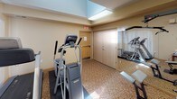 This fitness room is a dedicated exercise space designed to support a well-rounded cardio workout routine. The room features warm, neutral-toned walls paired with patterned carpeting that adds a touch of comfort to the functional environment. Large windows dressed with vertical blinds allow natural light to filter in, creating a bright and inviting atmosphere.

The space is equipped with a selection of cardio machines, including a treadmill, an elliptical trainer, and a recumbent stationary bike, all positioned thoughtfully throughout the room. Protective floor mats are placed beneath the equipment to preserve the flooring. A wall-mounted television is installed for entertainment during workouts, adding convenience for users.

A full-length wall mirror expands the perceived depth of the room and allows users to monitor their form while exercising. Overhead fluorescent lighting provides consistent illumination throughout the space. A small side table with a waste bin is also available for practical use.

The room is accessed through a solid wood-paneled door and features clear exit signage, ensuring safety and easy navigation. The elevated ceiling with a recessed upper section adds a sense of openness to the space. Overall, this fitness room offers a clean, functional, and comfortable environment for daily exercise.