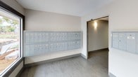 This mail area is a well-designed, single-floor communal space that serves as a functional hub for residents. The area is finished with a clean, neutral color palette featuring warm off-white walls, grey baseboards, and light tile flooring that runs consistently throughout, creating a cohesive and tidy atmosphere.

The space is anchored by wall-mounted mailbox units, which are arranged in organized rows of individual grey metal compartments. A smaller cluster of boxes occupies one section of the area, while a larger, multi-row bank of mailboxes is positioned along an adjacent wall, providing ample mail storage capacity for all residents. A doorway with wall-mounted lighting connects to the adjoining corridor, ensuring easy access.

Natural light plays a significant role in the space's ambiance. Large, dark-framed windows and a full glass entry door allow generous amounts of daylight to filter in, offering views of an outdoor landscaped area complete with wooden fencing and greenery. The textured ceiling adds a subtle architectural detail, while the overall layout remains open and uncluttered, prioritizing ease of movement and accessibility for daily use.